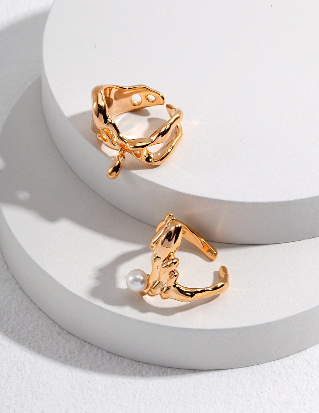 Set of two gold-plated rings including the Minimalist Teardrop Pendant Ring and a pearl-accented open band, styled on layered white platforms.