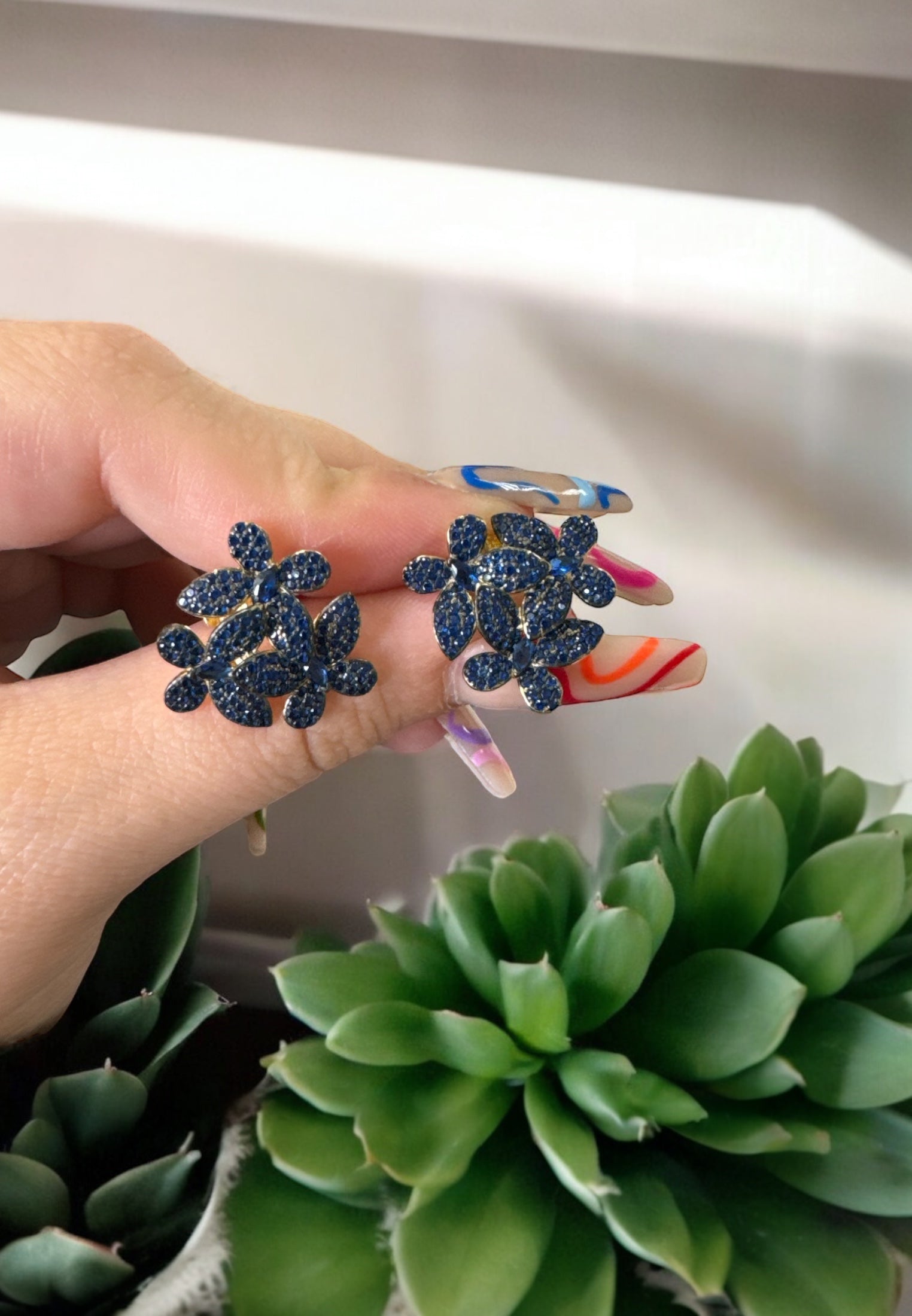 Hand holding sapphire blue flower stud earrings with gold base, displayed against green succulents for a natural aesthetic.