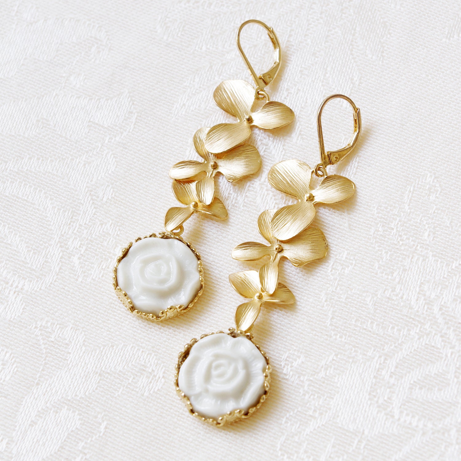 Elegant Porcelain Moonlight Rose Drop Earrings displayed on textured fabric, featuring golden triple leaves and handcrafted white rose details.