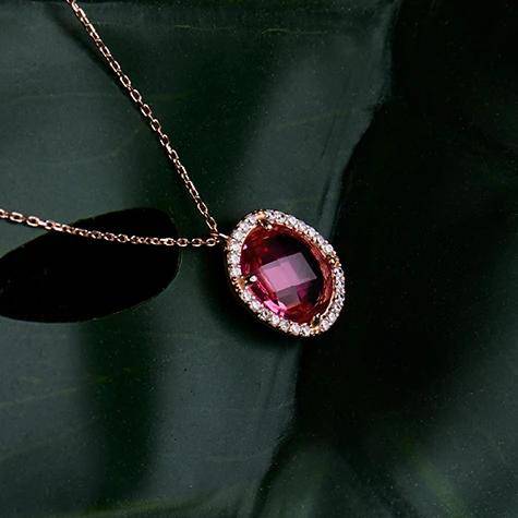Beatrice Oval Gemstone Pendant Necklace in rose gold with a pink tourmaline center, surrounded by sparkling crystals, displayed on a dark green background.
