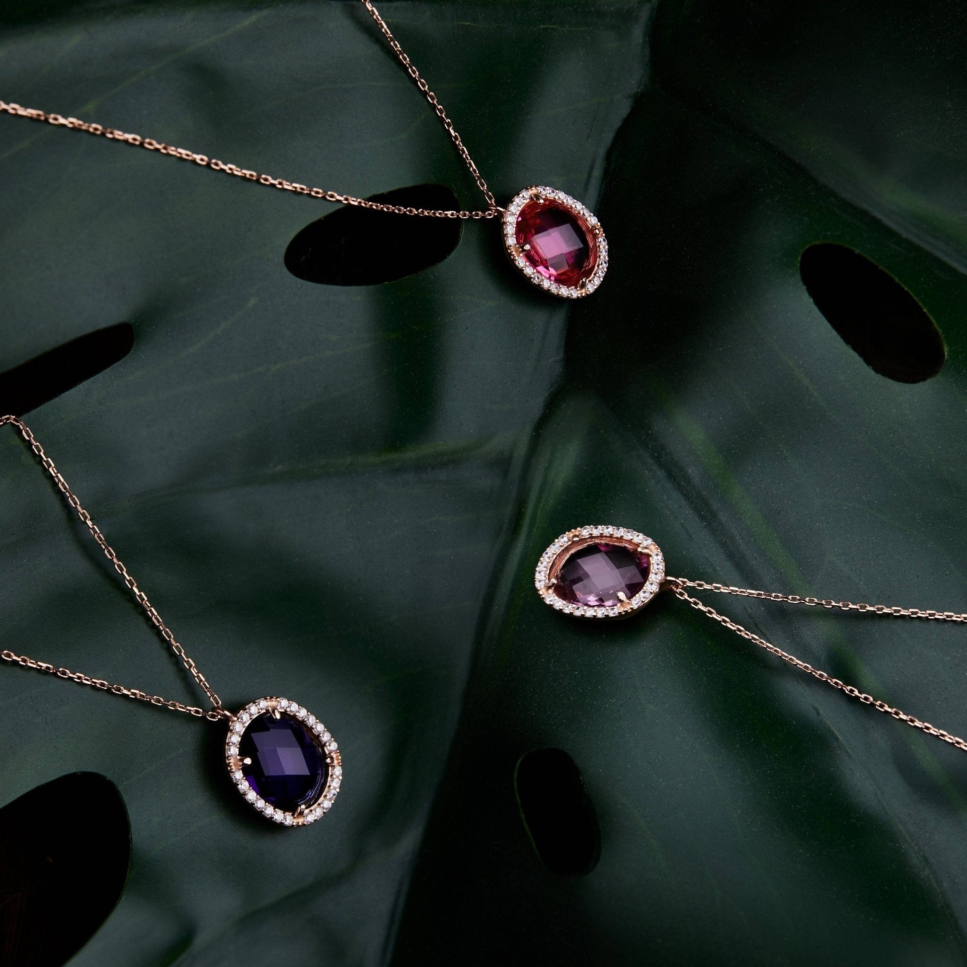 Beatrice Oval Gemstone Pendant Necklace Rose Gold Pink Tourmaline displayed on a dark green leaf with two other gemstone pendants. Elegant rose gold chain and halo setting enhance the pink tourmaline centerpiece.